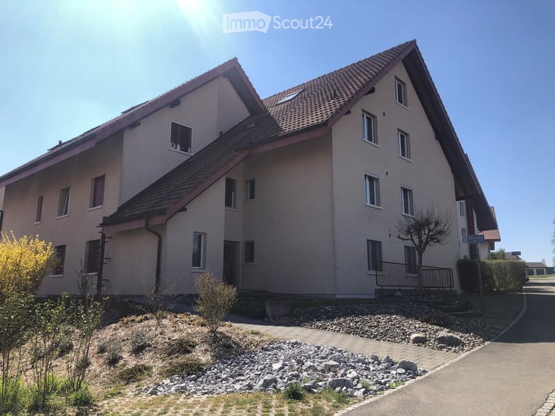 Two-story house with beige exterior, multiple windows, and a sloped roof. The house has a small balcony on the upper floor and is surrounded by a landscaped area with rocks and shrubs.