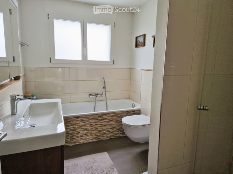 Modern bathroom with a white sink, bathtub, toilet, and a framed picture on the wall. The space features beige tiles and a wooden shelf with a small decorative item.