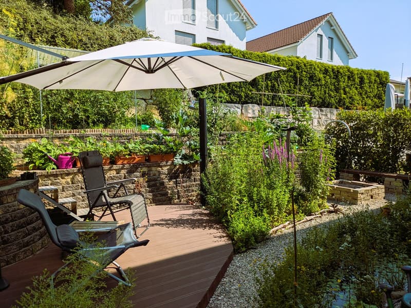 Garden with wooden deck, lounge chairs, umbrella, plants, and a small water feature.