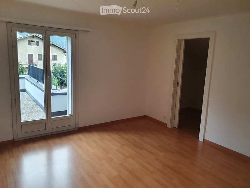 Empty room with wooden flooring, large sliding glass doors leading to a balcony, and a visible adjacent dark room.