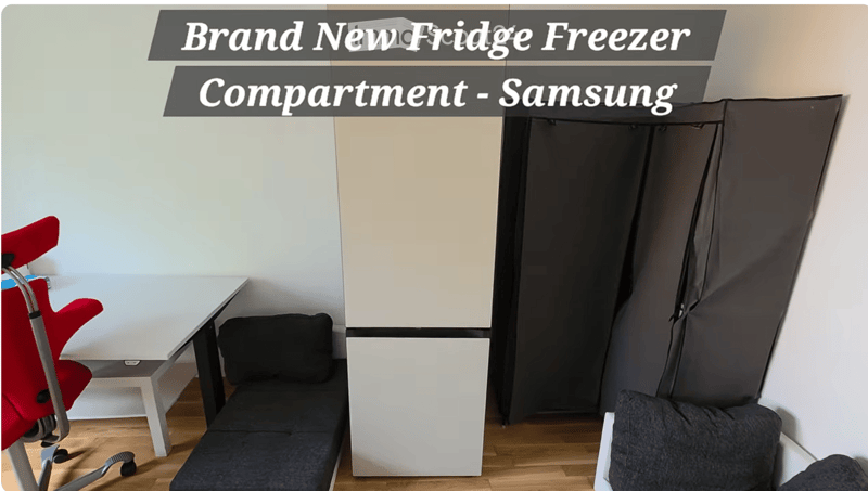A compact, furnished room with a sofa, chair, small table, and a Samsung fridge freezer. The room features light-colored walls and wooden flooring.