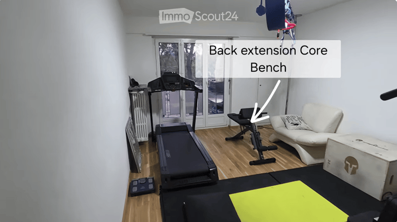A well-lit room with wooden flooring, featuring a white sofa, a bench, a treadmill, an exercise bike, a scale, and a storage box.