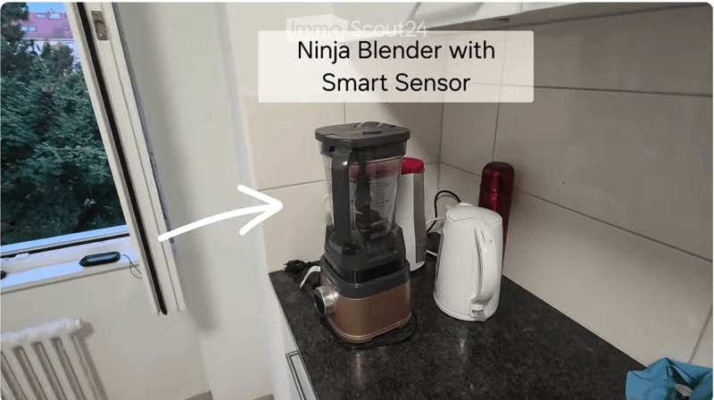 Modern kitchen counter with a Ninja Blender with Smart Sensor, a white kettle, and a cup. The counter is dark, and there is a window with a view of greenery.