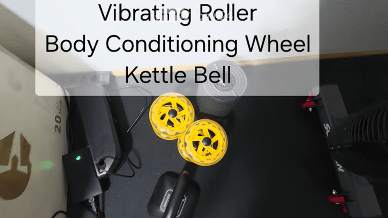 A collection of fitness equipment including a body conditioning wheel, kettle bell, and vibrating roller placed on a black mat.