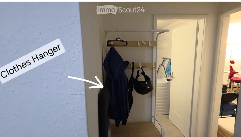 A narrow hallway with a clothes hanger on the wall, leading to an open door revealing a room with furniture.