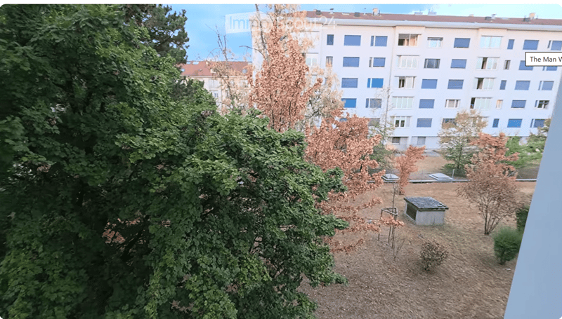 View of a garden area with trees, shrubs, and a small structure, with a large residential building in the background.