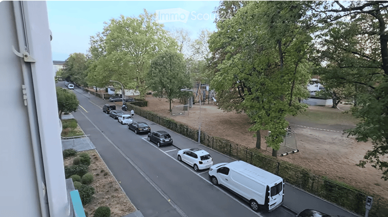 Balcony view shows a quiet residential street with parked cars, trees, and a small park area with playground equipment.