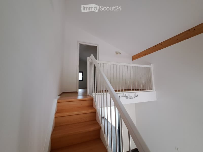 A modern staircase with wooden steps and a white metal railing, leading to an upper level with white walls and a wooden ceiling beam.