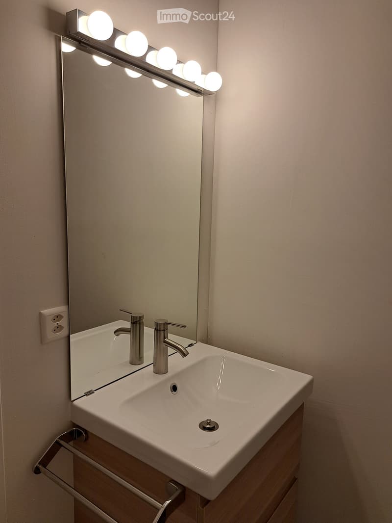 Modern bathroom with a white sink, dual faucets, a large mirror with integrated LED lighting, and a wooden cabinet.