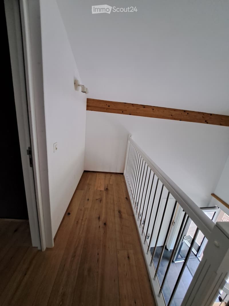 A narrow hallway with wooden flooring, white walls, and a metal railing leading to an open area. The hallway features a wooden trim along the wall and a light switch on the left side.