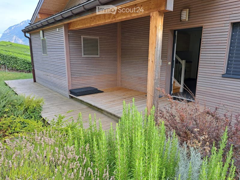 A modern wooden house with a raised wooden deck, surrounded by greenery and a scenic mountain view in the background.