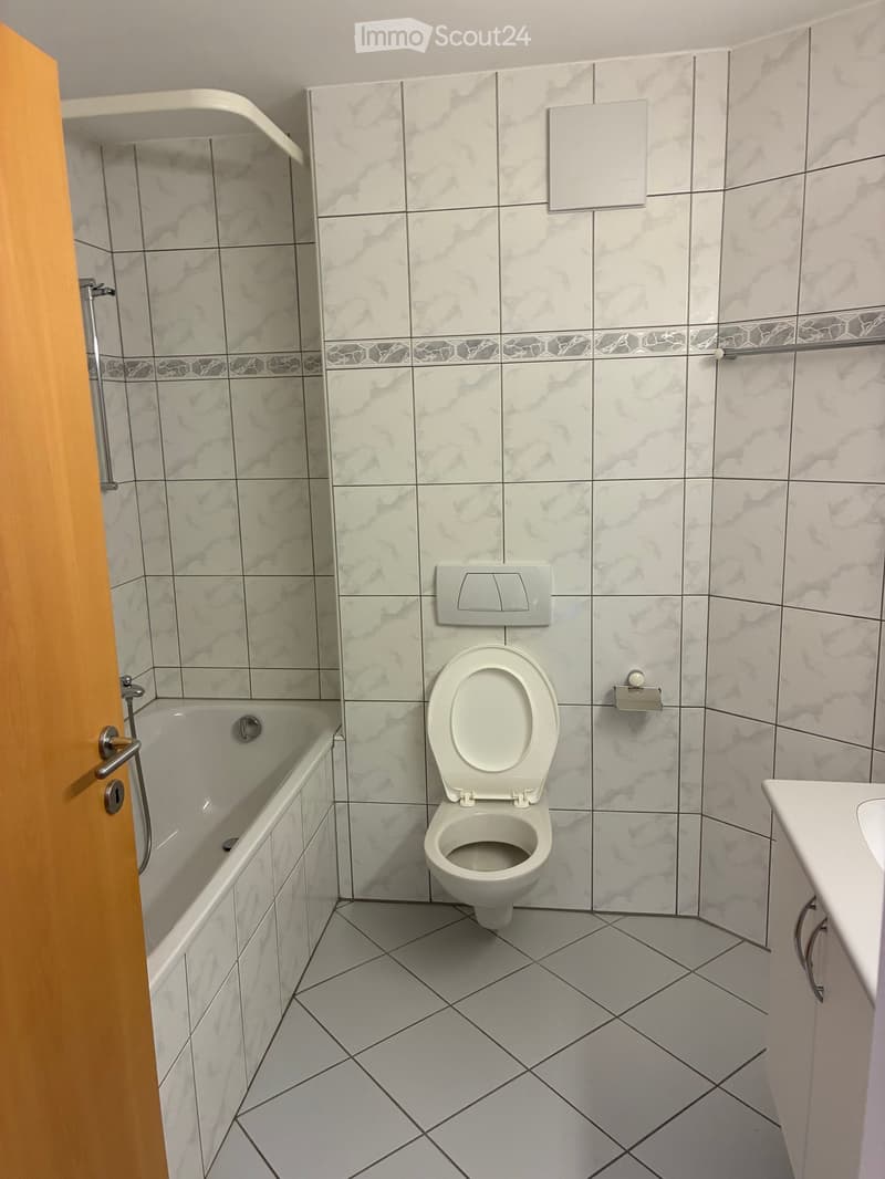 White tiled walls and floor, bathtub, shower, toilet, sink, and a wall-mounted soap dispenser