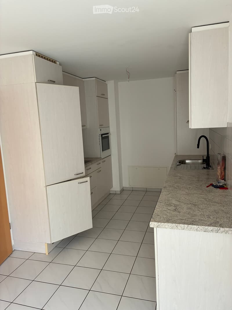 A small kitchen with white tiled flooring, white cabinetry, and a granite countertop. It includes an oven and a stove, a sink with a faucet, and a small area for a dishwasher.