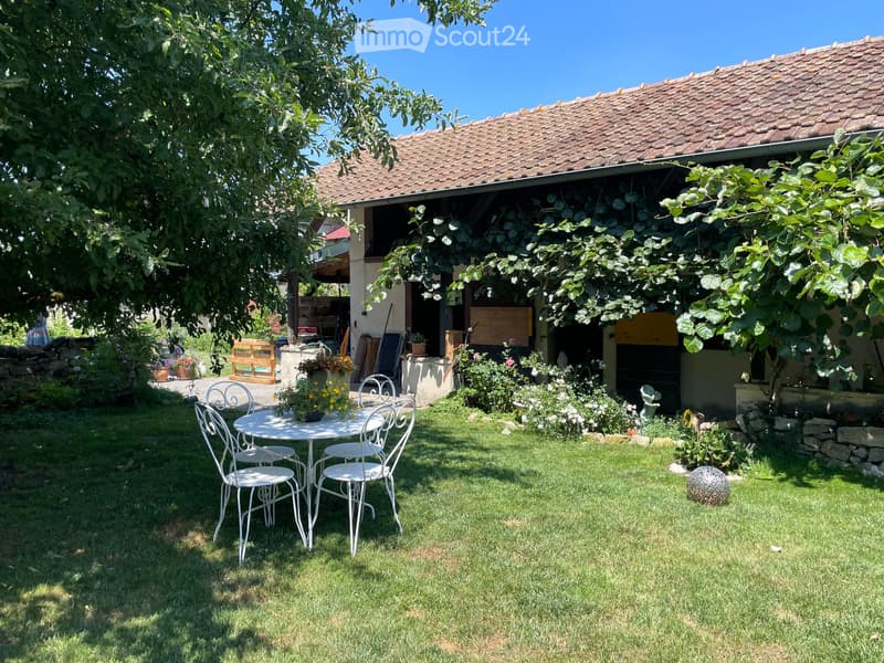 Garden area with a white metal table and chairs, surrounded by greenery and a house with a tiled roof.