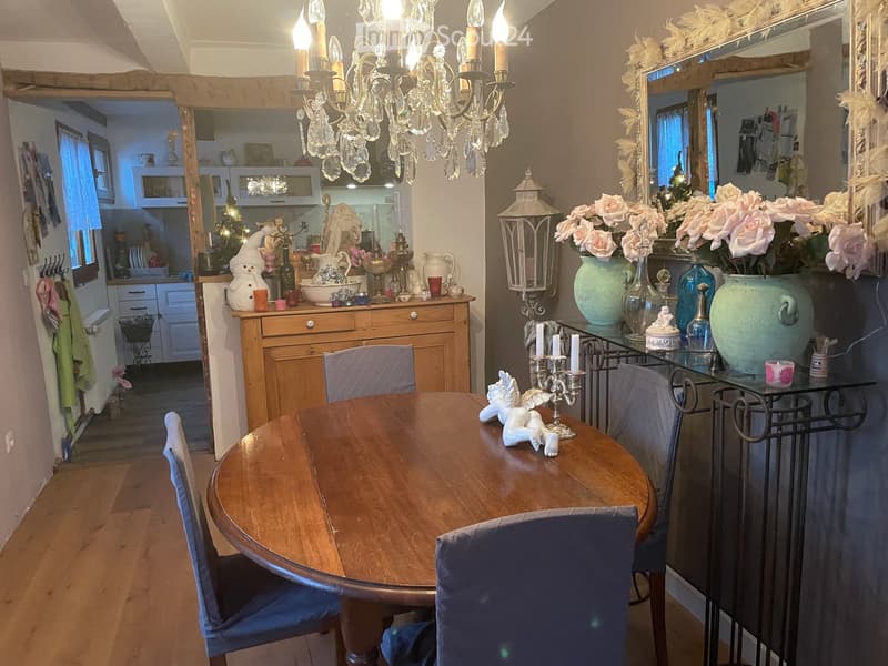 A cozy dining area with a round wooden table, four chairs, a chandelier, and decorative vases on a side table. The background shows a kitchen area with cabinets and a countertop.