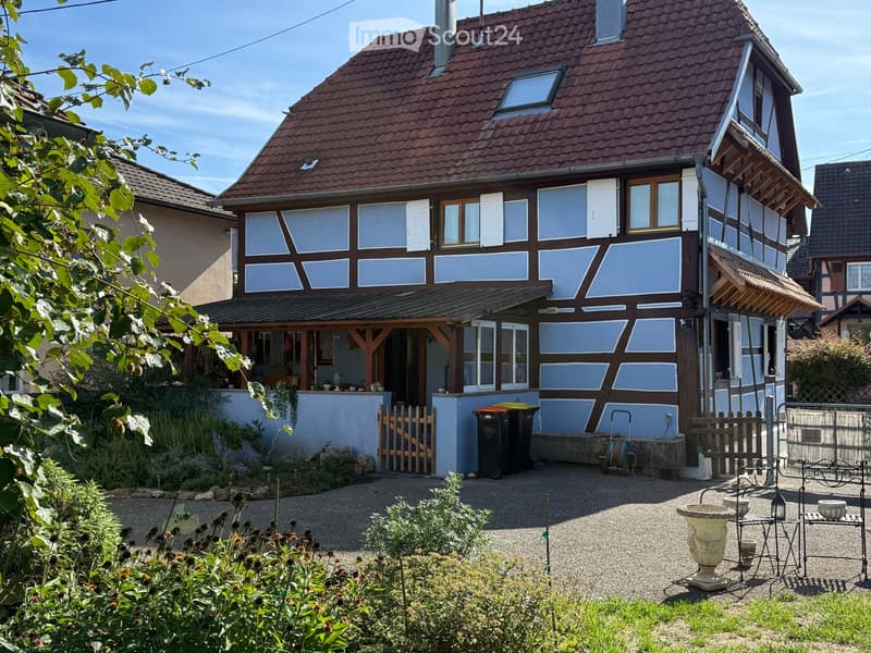 Traditional two-story house with a red-tiled roof, light blue and brown facade, and a covered porch area. The garden features gravel pathways, plants, a decorative urn, and a small wooden fence.