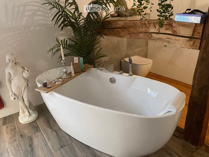 Modern bathroom with a large freestanding bathtub, toilet, wooden shelf with candles and decorative items, and a statue on the floor.