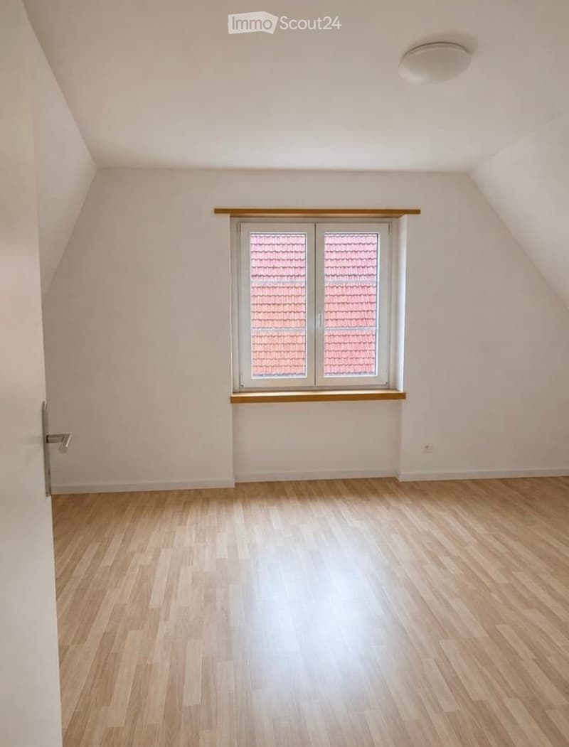 Empty attic room with wooden flooring, white walls, and a large window with a wooden sill. The ceiling has a sloped design typical of attic spaces.