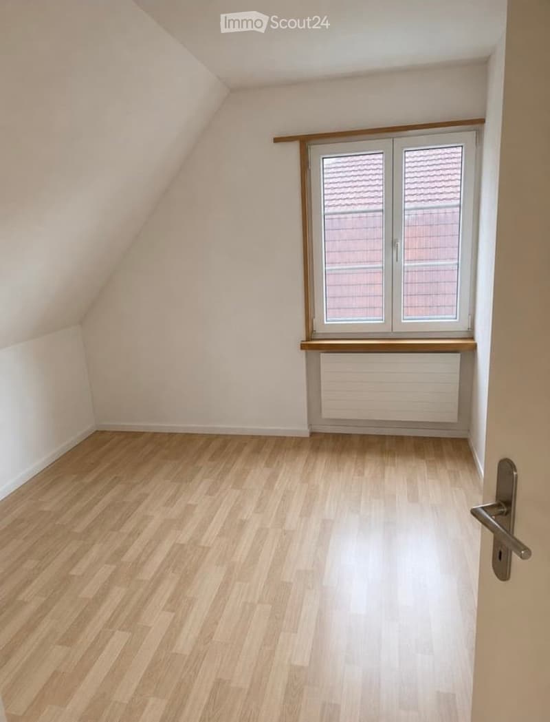 Empty attic room with wooden flooring, white walls, and a large window with a radiator below. The room has a slanted ceiling typical of attic spaces.