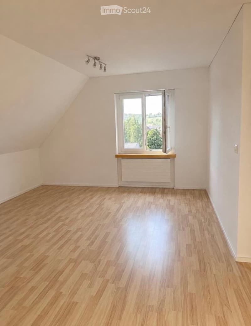 Empty room with wooden flooring, white walls, and a large window with a wooden sill. The room has a slanted ceiling and built-in lighting fixtures.
