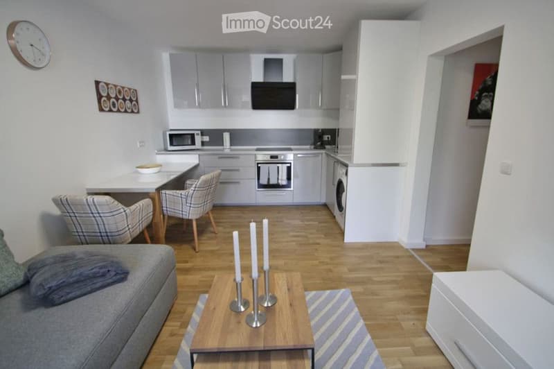Modern open-plan living and dining area with a grey sofa, coffee table, dining table with chairs, and a fully equipped kitchen featuring a microwave, oven, and washing machine.