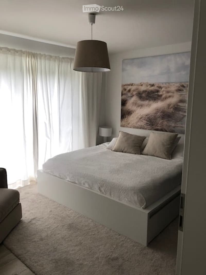 Modern bedroom with a large bed, neutral-colored bedding, two throw pillows, a hanging pendant light, a large window with white curtains, a wall-mounted painting, and a small side chair.