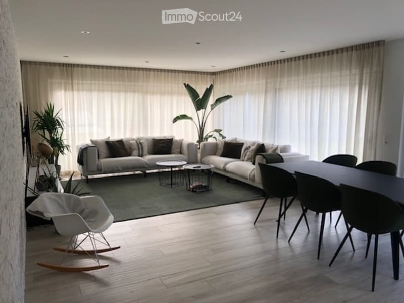Modern living room with a grey sectional sofa, coffee tables, a white rocking chair, and a dining table with chairs. Large windows with sheer curtains and indoor plants add to the decor.