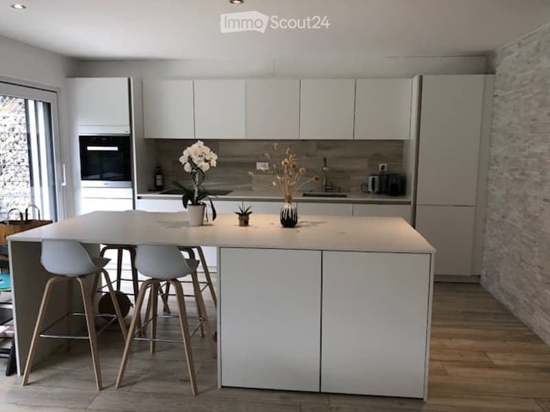 Modern kitchen with white cabinets, a central island, and four stools. Features include a built-in oven, refrigerator, and decorative vases on the island.