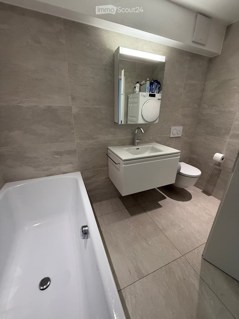 Modern bathroom with a bathtub, sink, toilet, and a mirrored cabinet above the sink. The walls and floor are tiled in a light, neutral color.