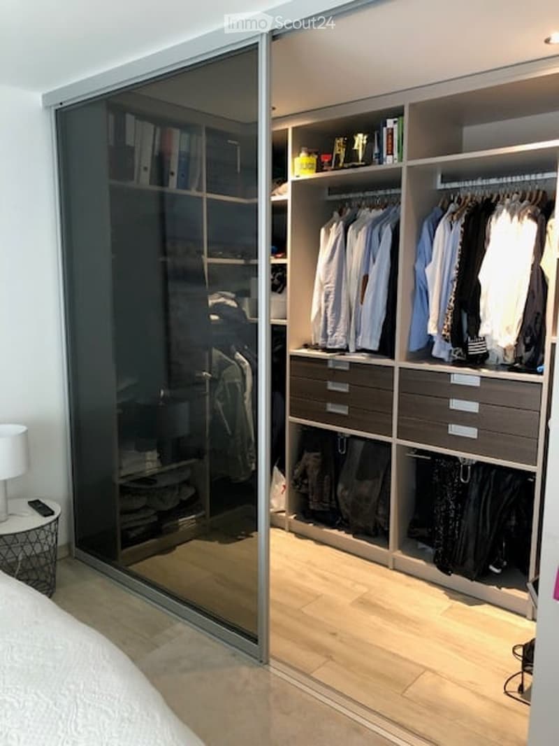 Modern bedroom with a large, mirrored wardrobe, neatly organized with clothes and accessories. A bed is partially visible, and a small table with a lamp is placed near the wardrobe.