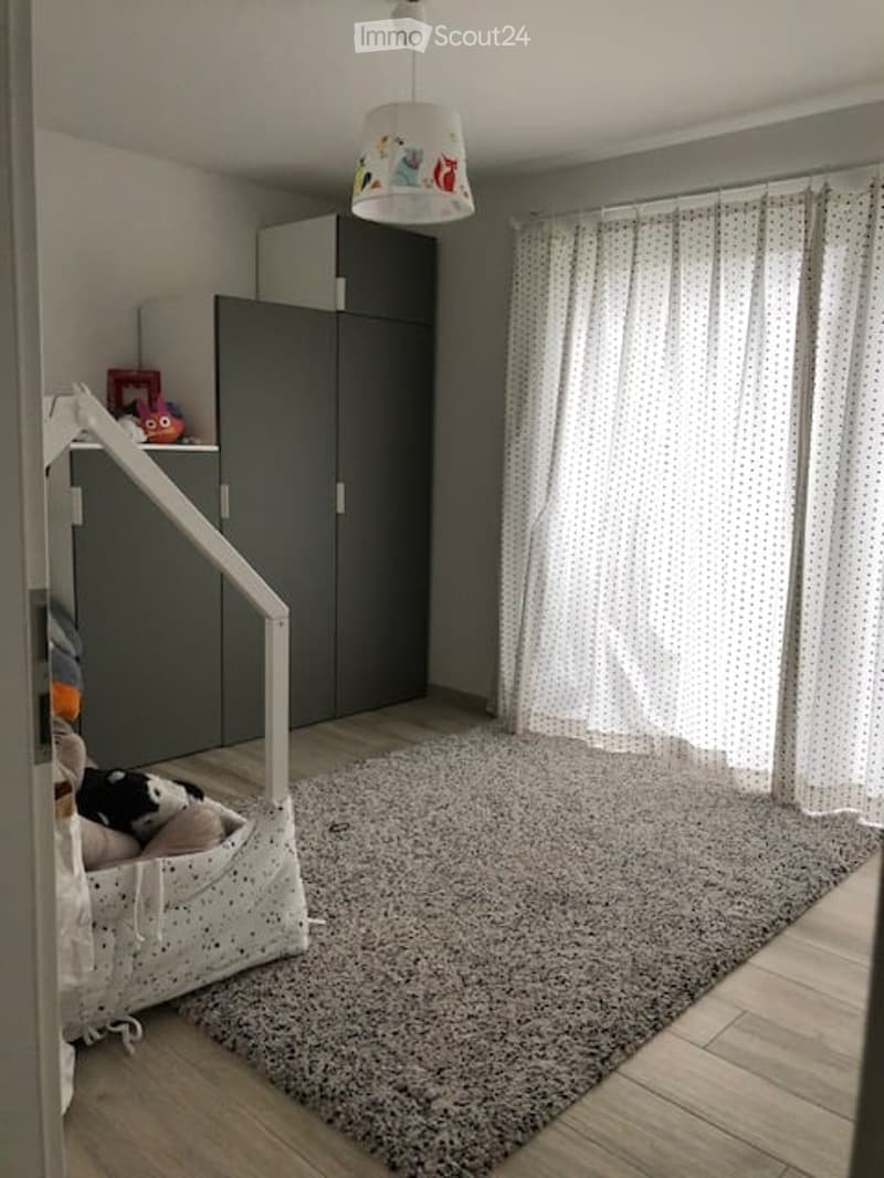 A well-lit children's room with a grey wardrobe, a large window with white curtains, a soft grey rug, and a toy basket with stuffed animals.