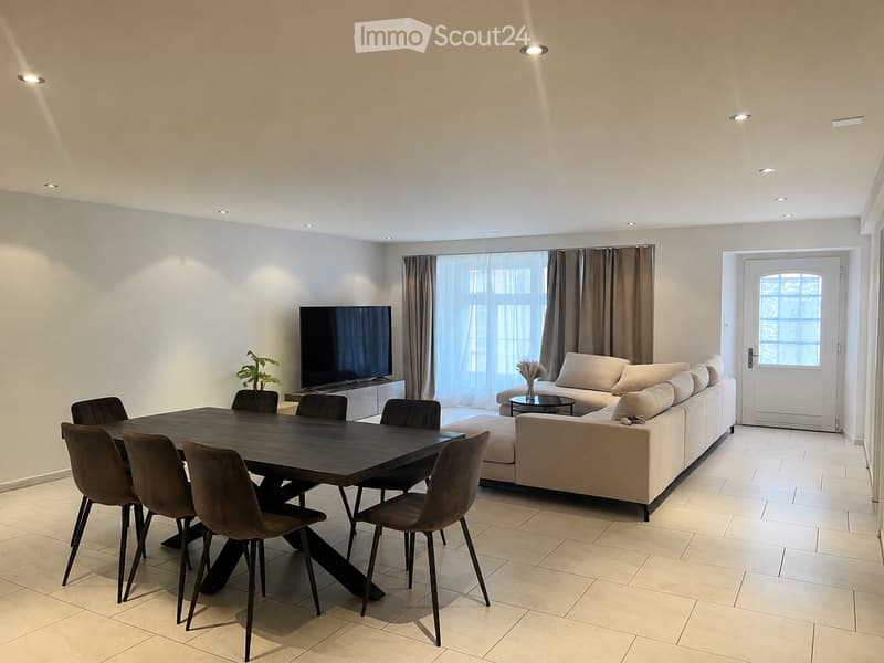 Modern living and dining area with a large sectional sofa, coffee table, flat-screen TV, dining table with six chairs, and a small side table with a vase.