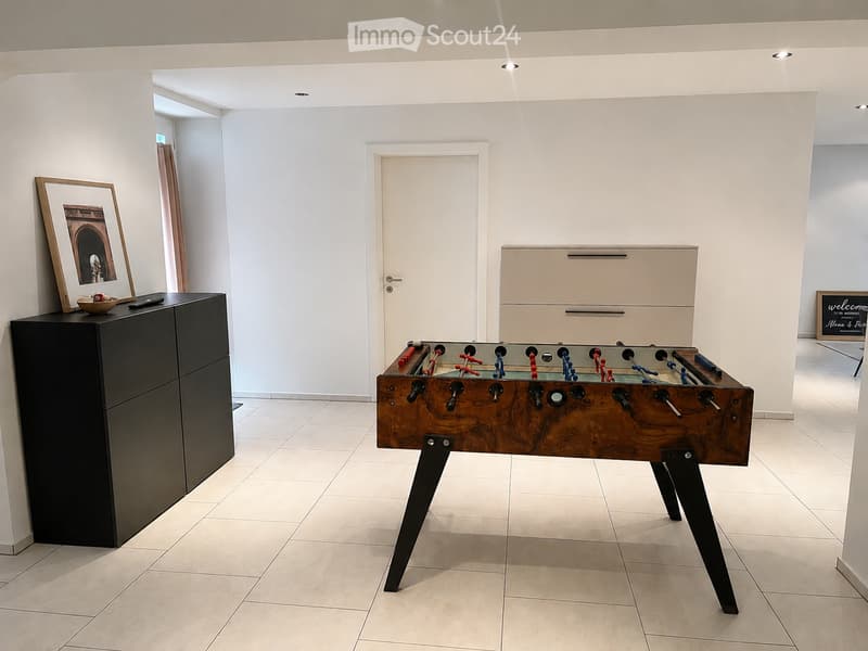 A modern, minimalistic room with a foosball table as the central feature, a black cabinet with a picture frame and bowl on top, and a white door in the background.