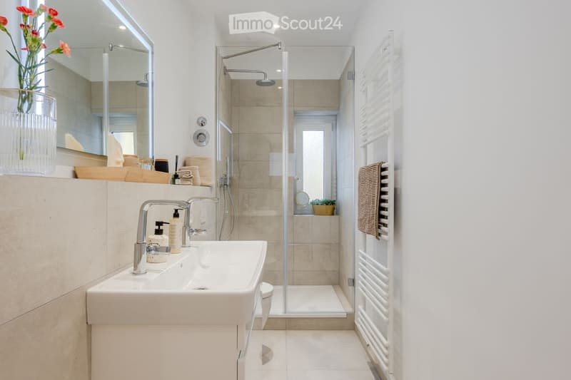 Modern bathroom with a double sink, large mirror, glass-enclosed shower, towel rack, and decorative vase with flowers.