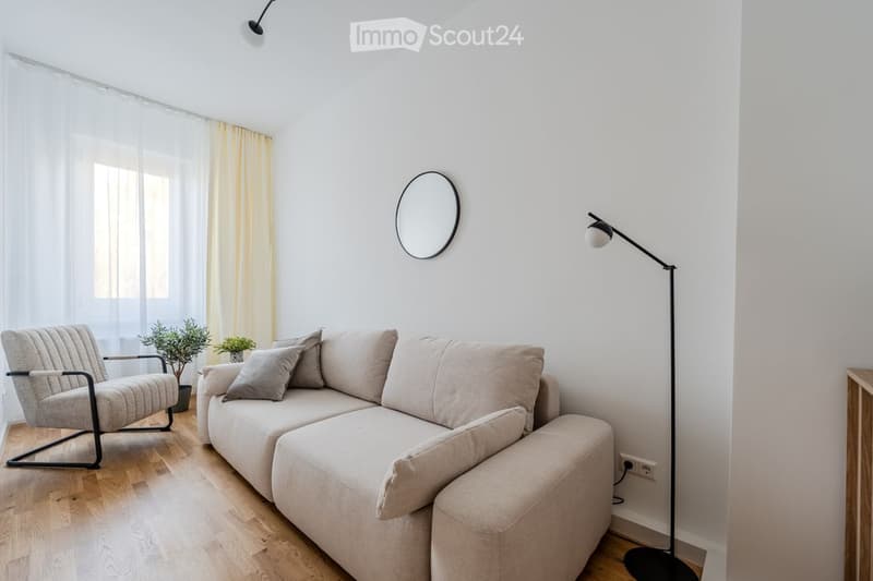 Modern living room with a light grey sectional sofa, matching armchair, floor lamp, round wall mirror, and small potted plants. The room features light wooden flooring and large windows with sheer curtains.