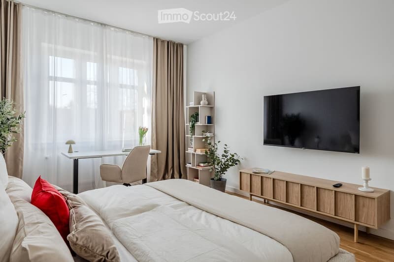 Modern bedroom with a large bed, nightstand, desk, chair, lamp, shelf, plant, and television mounted on the wall.