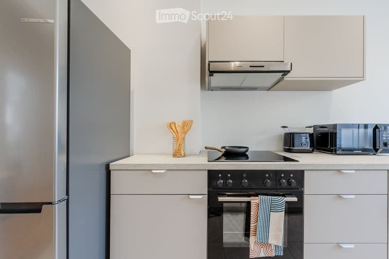 Modern kitchen with stainless steel appliances, white cabinets, and a light-colored countertop. Includes a refrigerator, oven, microwave, toaster, frying pan, wooden utensils, and kitchen towels.