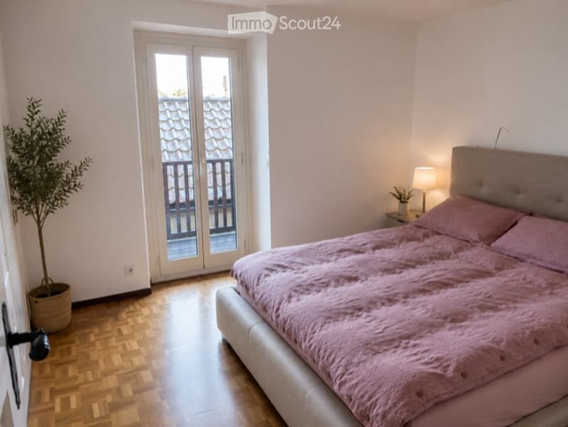Bright bedroom with wooden flooring, a large bed with pink bedding, a nightstand with a lamp, and a potted plant near the balcony door.