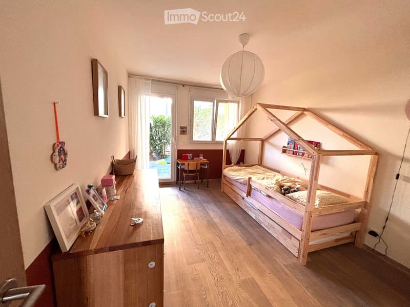 Bright bedroom with wooden floor, large window, and a unique wooden bed shaped like a house. Includes a desk, chair, mirror, picture frames, and a wicker basket.