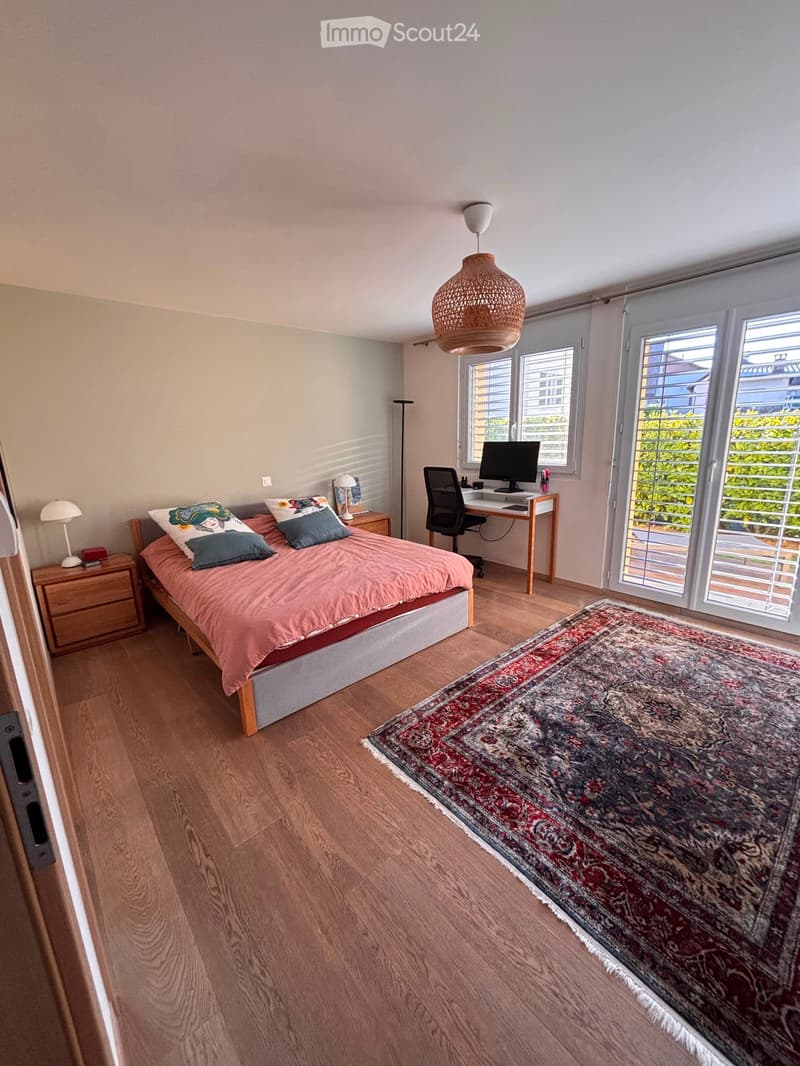 Bright bedroom with wooden flooring, large windows, a bed with pink bedding, a nightstand, a desk with a computer, and a patterned rug.