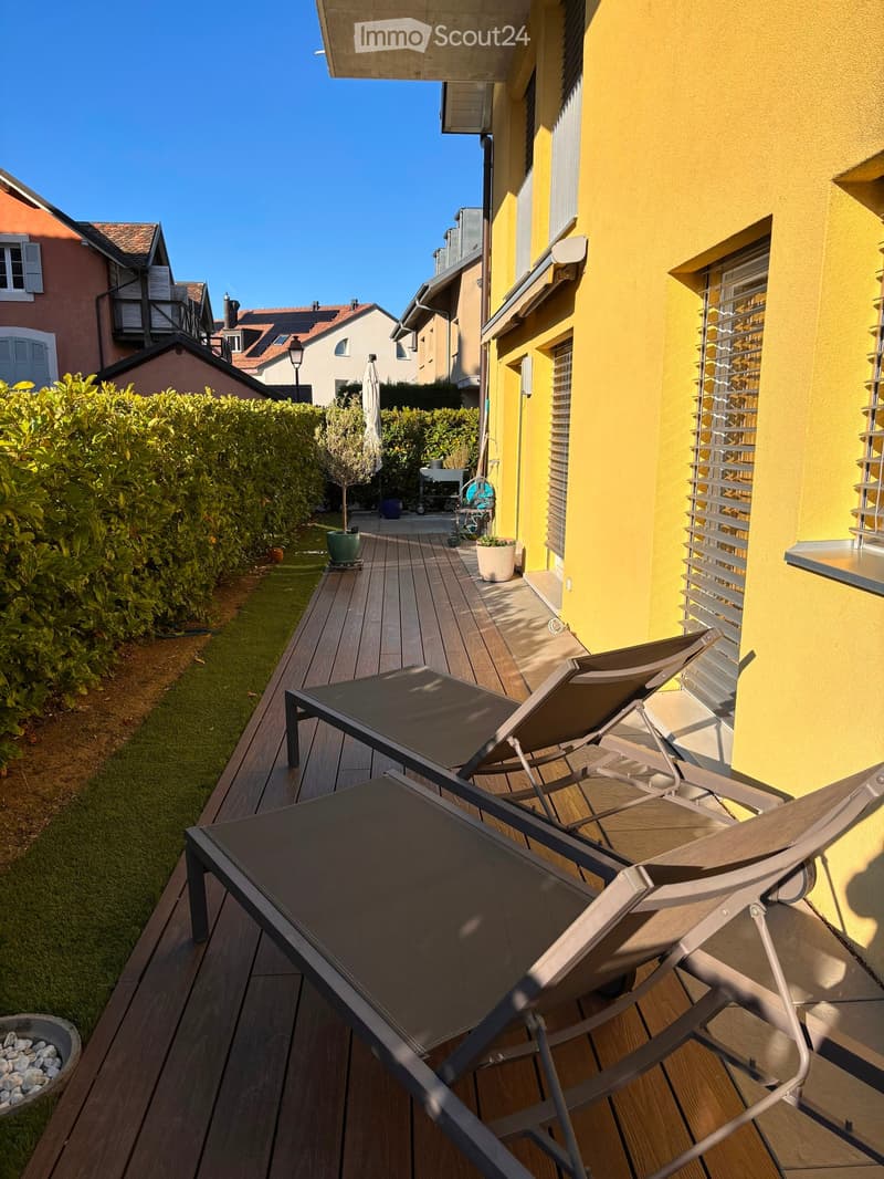 Balcony with wooden decking, table, and two lounge chairs, adjacent to a yellow building with shutters.