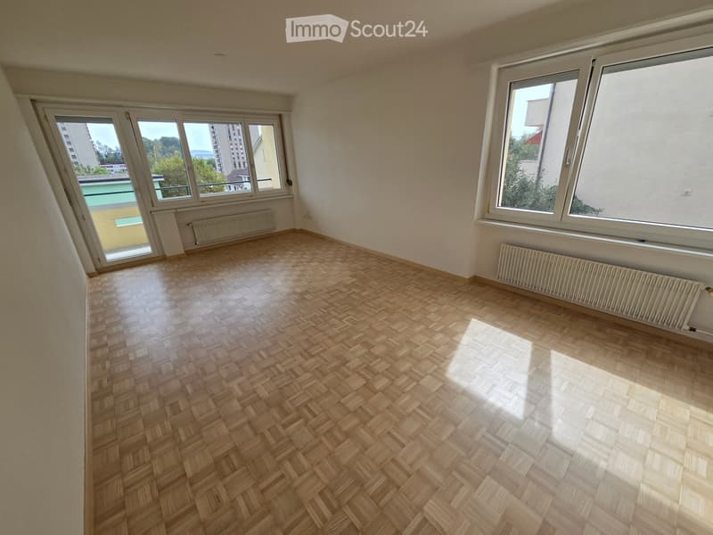 Empty room with wooden herringbone flooring, large windows, radiators, and a view of a building and greenery outside.