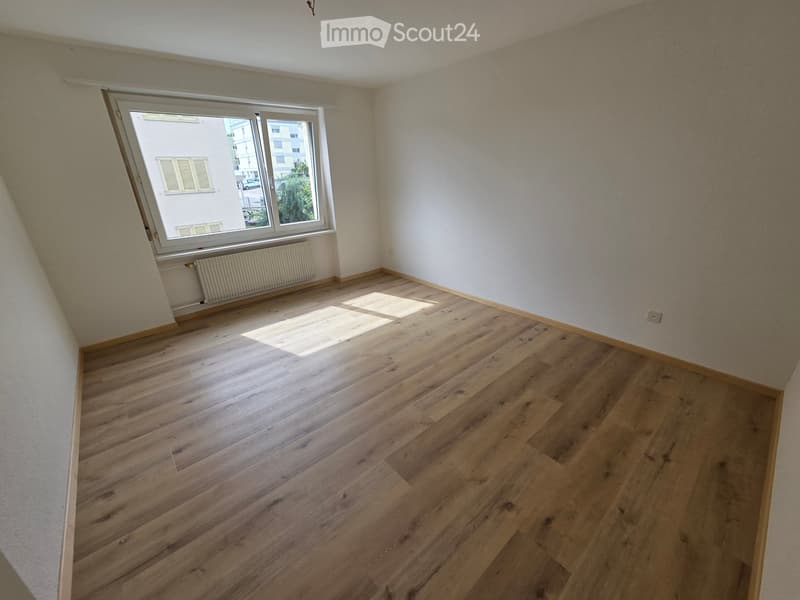 Empty room with wooden flooring, white walls, and a large window letting in natural light. The room has a radiator below the window and a visible electrical outlet on the wall.