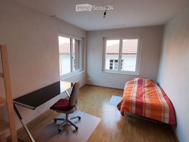 A small, furnished bedroom with a single bed, a desk, and a chair. The room has wooden flooring and large windows offering a view of the roof.