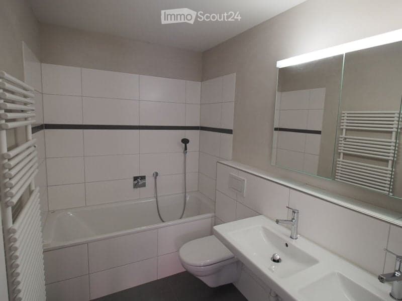 Modern bathroom with white tiled walls, a bathtub, toilet, sink, and a large mirror with built-in lighting. A white radiator is mounted on the wall.