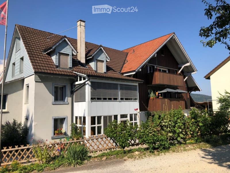 Two-story house with a combination of brown and red tile roofs, light grey walls, multiple windows, and a wooden balcony with outdoor furniture.