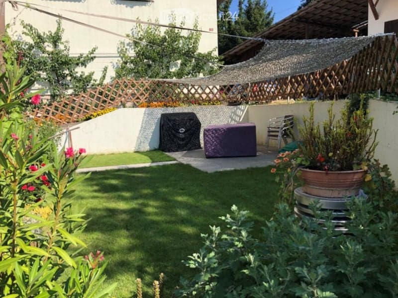 A small, well-maintained garden with artificial grass, a wooden lattice fence, potted plants, and outdoor furniture including a covered grill and a small table and chairs.