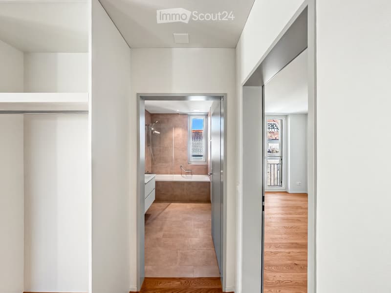 A modern, minimalist hallway with white walls and wooden flooring, leading to a bathroom and another room with a balcony view.
