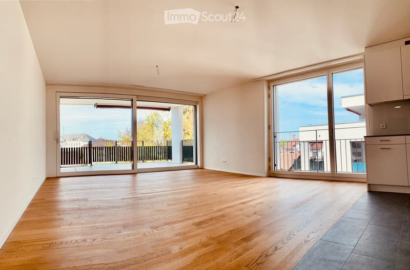 Empty room with wooden flooring, large sliding glass doors leading to a balcony with a view of the city and trees.