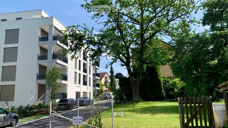 Modern multi-story residential building with balconies, surrounded by greenery and parked cars.
